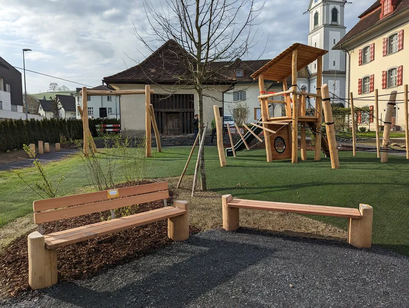 Der Generationenpark vom Boden aus: Holzbänke zum Sitzen, dahinter der Spielturm, im Hintergrund Kirche und Häuser von Pfaffnau.